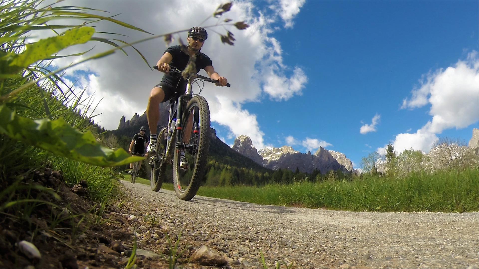 Ciclisti su un sentiero di montagna, cielo azzurro e montagne sullo sfondo.
