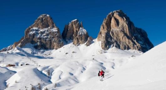 Due sciatori attraversano un paesaggio innevato con montagne maestose sullo sfondo.