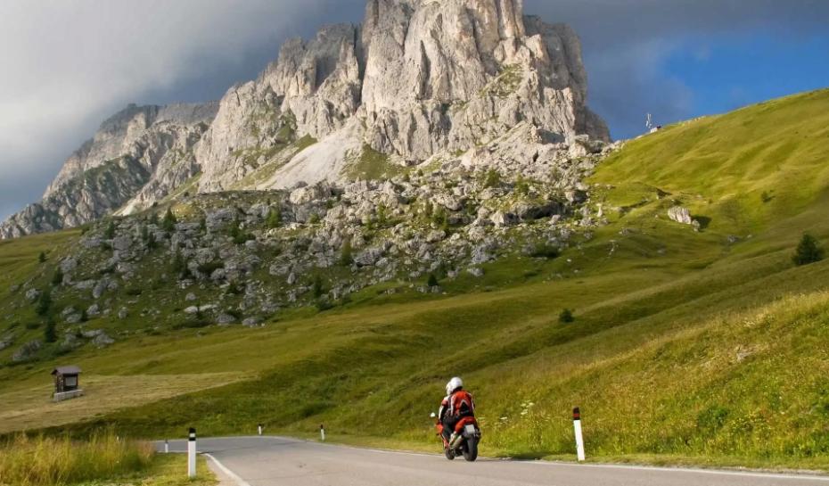 Motociclista percorre strada di montagna con imponente roccia sullo sfondo, cielo parzialmente nuvoloso.